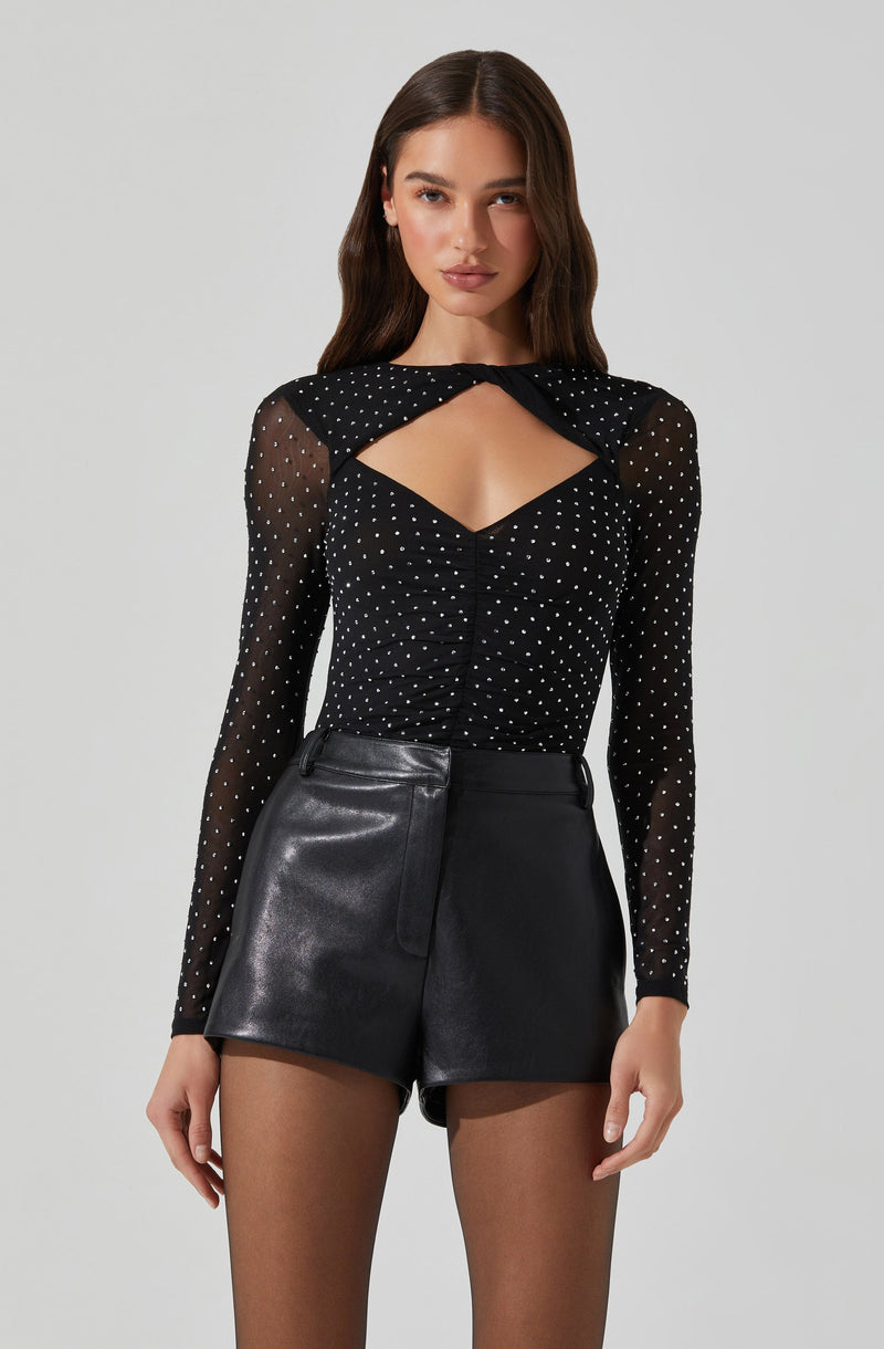 A woman is wearing a black, long-sleeved, sheer top with white polka dots and a distinctive cutout design at the chest. The top has a black bow detail at the neckline. She pairs it with high-waisted black leather shorts.