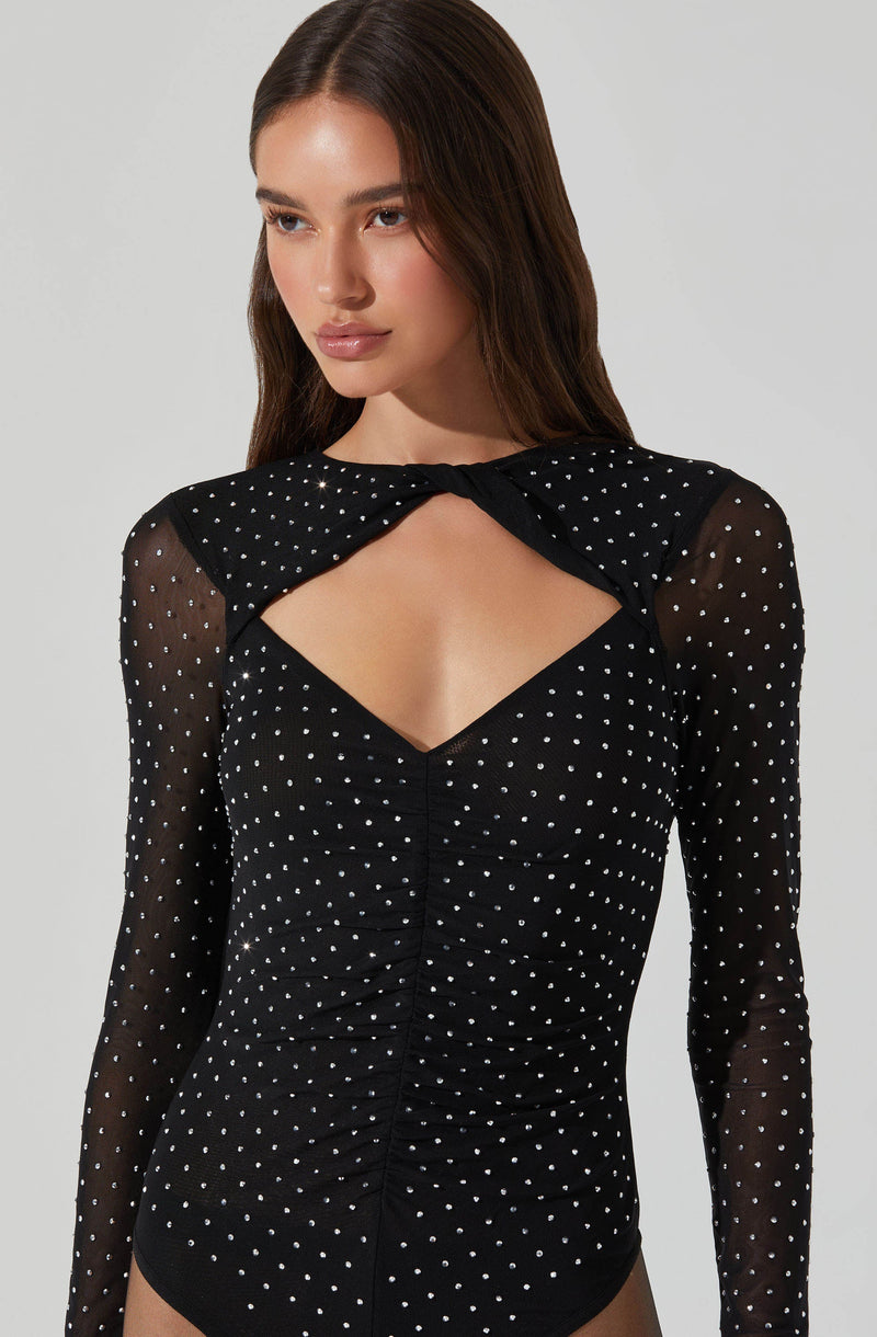 A woman is wearing a black long-sleeved bodysuit with white polka dots. The bodysuit features a sheer fabric on the sleeves and shoulders, a central ruched detail, and a distinctive cut-out at the chest area.