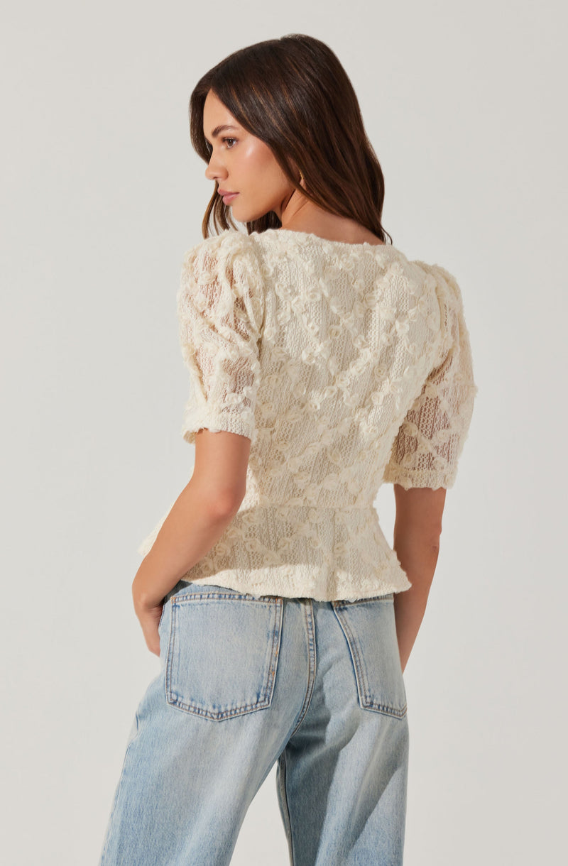 A woman is wearing a cream-colored, lace blouse with puffed short sleeves and a peplum hem. She is paired it with light-wash, high-waisted jeans.