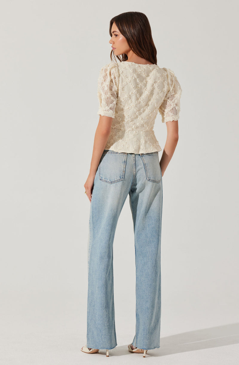 A woman is wearing a cream-colored lace top with short, puffed sleeves and a slightly flared hem. She pairs it with high-waisted, light-wash jeans that have a loose fit and prominent back pockets. She is also wearing light-colored high-heeled shoes.