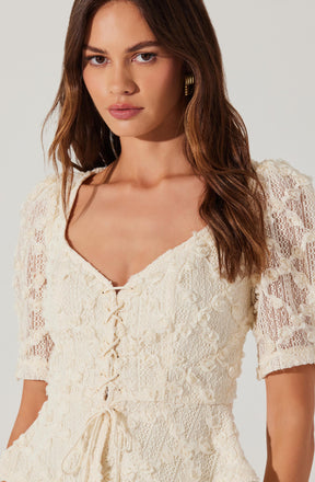 A woman is wearing a cream-colored, short-sleeved lace dress with a V-neckline. The dress features intricate lace detailing and a front tie closure with a bow at the waist.