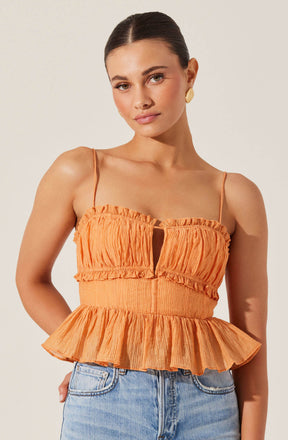 A woman is wearing a sleeveless orange top with thin spaghetti straps. The top features a gathered, ruched fabric with a keyhole opening in the center and a ruffled hem that flares out at the waist. She has paired the top with light blue high-waisted jeans.