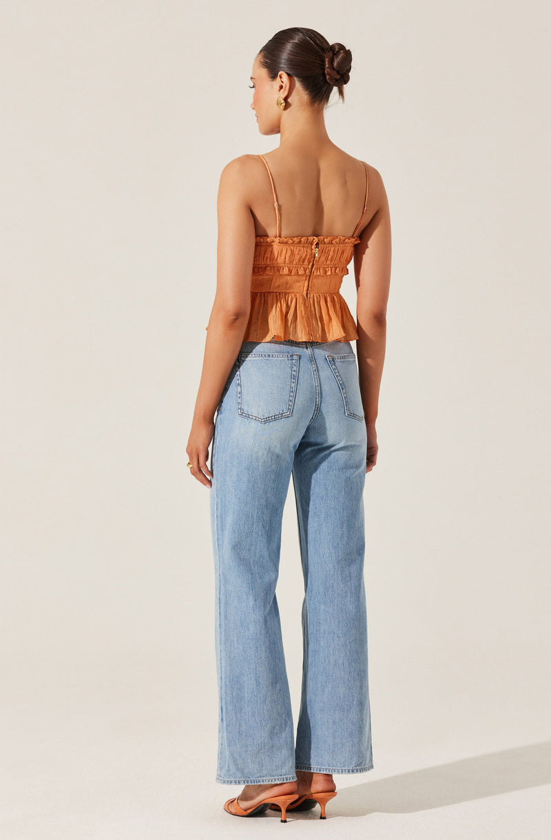 A woman is wearing a sleeveless, orange, ruffled top with spaghetti straps and a cinched waist. She pairs it with high-waisted, light-wash, wide-leg jeans and tan heeled sandals. She has tied back her hair in a bun and is wearing large gold hoop earrings.