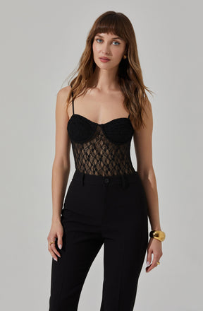 A woman is wearing a black lace bustier top with thin spaghetti straps and a sheer, patterned lace design. She pairs it with high-waisted black pants. She accessorizes with a chunky gold bracelet on her left wrist and a gold ring on her right hand.