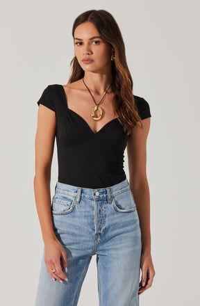 A woman is wearing a fitted black top with cap sleeves and a sweetheart neckline. She pairs it with high-waisted, light-wash jeans. She accessorizes with a large gold circular necklace and matching gold hoop earrings.