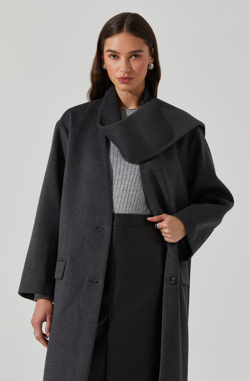 A woman is wearing a dark gray, oversized wool coat with a wide collar that drapes across her shoulder. Underneath, she has on a light gray, ribbed sweater. She is also wearing black high-waisted pants. She accessorizes with pearl stud earrings and a ring on her left hand.