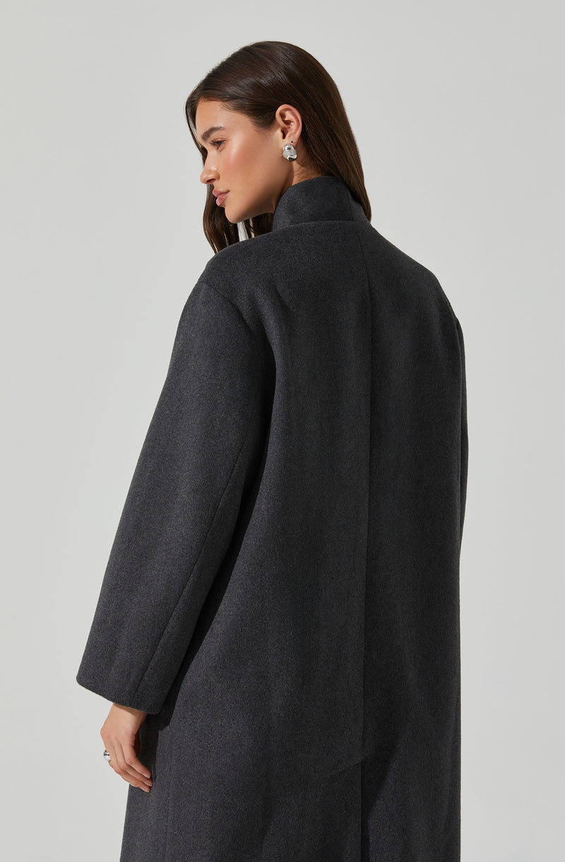 A woman is wearing a long, dark gray coat with a high collar. The coat appears to be made of a wool or wool-blend fabric, and it has a loose, relaxed fit. She is also wearing a pair of large, round earrings.