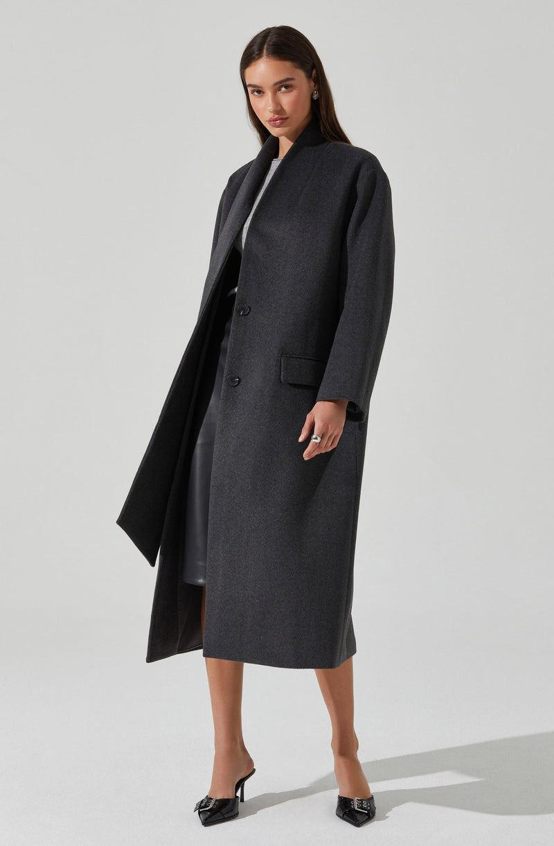 A woman is wearing a long, dark gray overcoat with a smooth, structured fit. The coat features a collarless neckline, a three-button front closure, and a flap pocket on the right side. She is also wearing black high-heeled shoes with pointed toes and decorative embellishments.
