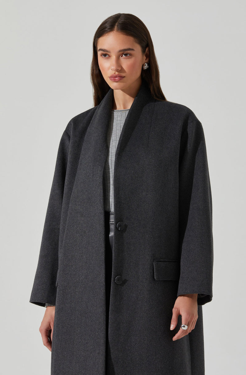 A woman is wearing a long, dark gray overcoat with a wide collar and a single button closure. The coat features a pocket on the front right side. The sleeves are slightly loose, and the overall style is sleek and minimalistic. She is also dressed in a light-colored top underneath.