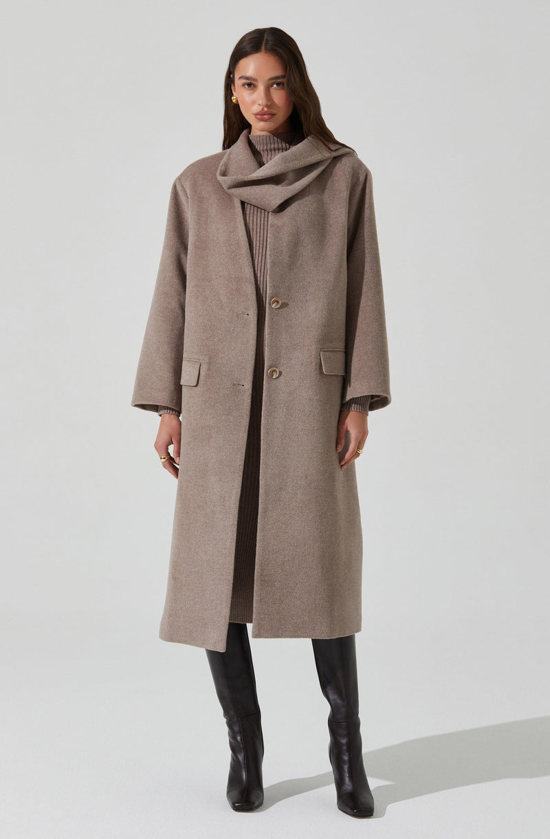 A woman is wearing a long, beige overcoat with a high collar and two large buttons near the waist. The coat features front pockets with flaps, and the sleeves are slightly loose. She pairs it with a matching beige turtleneck sweater underneath and black knee-high boots.