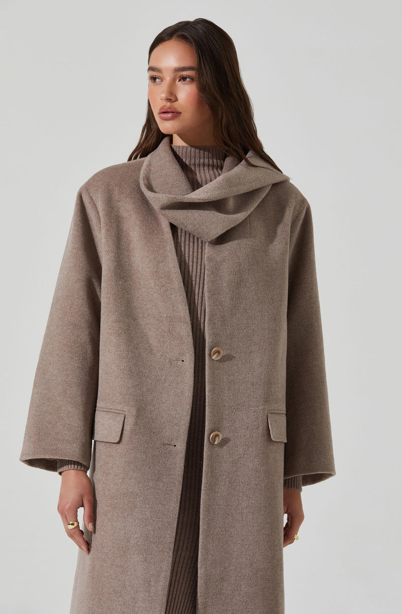 A woman is wearing a long, taupe-colored overcoat with a classic design, featuring two large front buttons and two flap pockets on either side. Underneath, she has on a ribbed, taupe-colored sweater with a high neckline. The coat has wide sleeves and a relaxed fit, complemented by a scarf or collar detail that drapes around her neck.