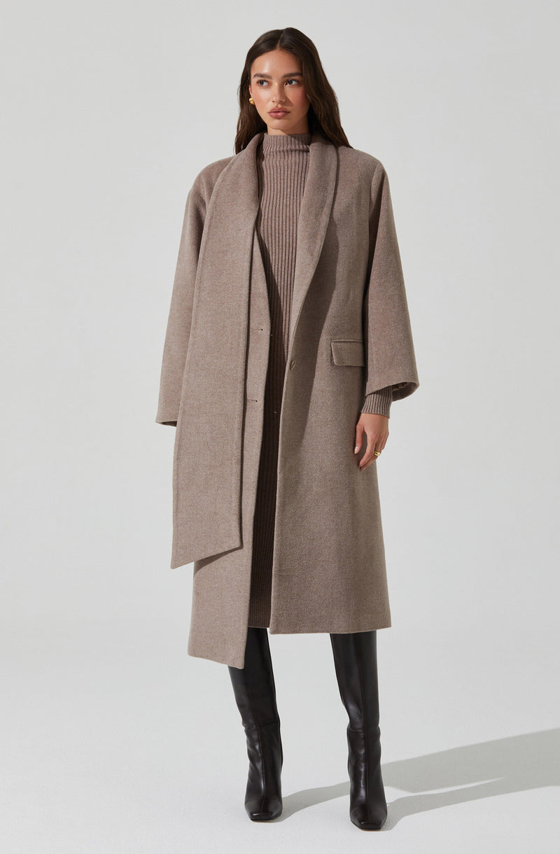 A woman is wearing a long, light beige overcoat with a tailored fit and wide lapels. Underneath, she has on a ribbed, high-neck sweater in a similar neutral tone. She pairs the outfit with black, high-shaft leather boots.