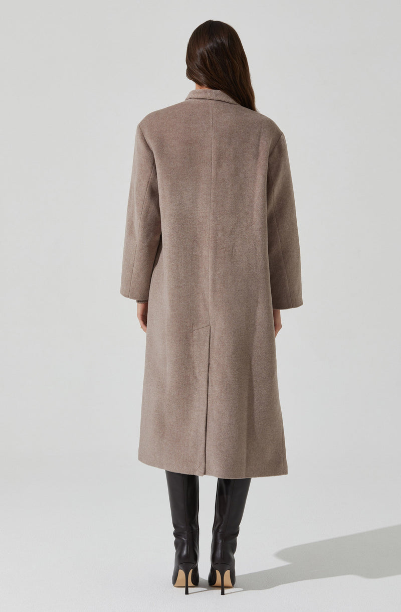A woman is wearing a long, beige overcoat that extends to her ankles. The coat features a tailored fit with a slightly structured collar and long sleeves. She is also wearing tall, black leather high-heeled boots. Her hair is down, and she is facing away from the camera against a plain, light-colored background.