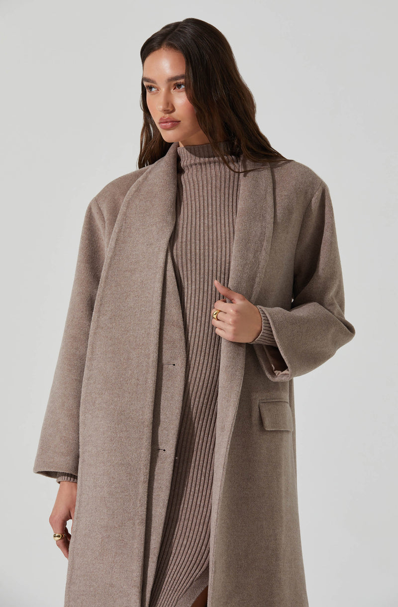 A woman is wearing a long, beige overcoat with a subtle texture and a wide lapel. Underneath, she has on a fitted, ribbed, high-neck sweater in a matching taupe color.