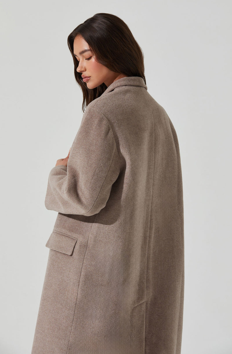 A woman is wearing a long, beige overcoat made of a soft, wool-like material. The coat features a collar and a vent at the back, and has a pocket with a flap on the side.