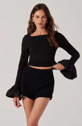 A woman is wearing a black long-sleeve crop top with a boat neckline and exaggerated ruffle cuffs at the wrists, paired with high-waisted black shorts.