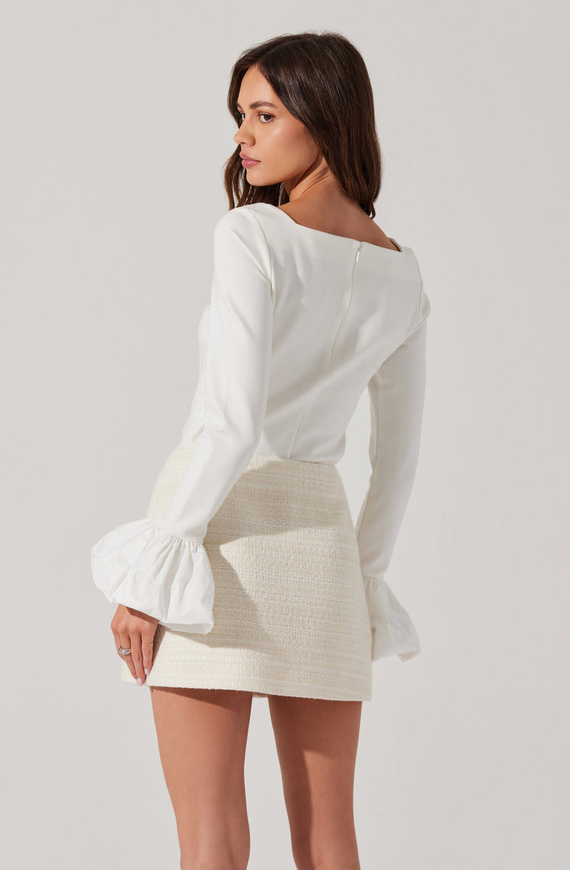 A woman is wearing a white long-sleeve blouse with a square neckline and dramatic ruffled cuffs, paired with a high-waisted cream-colored textured mini skirt.