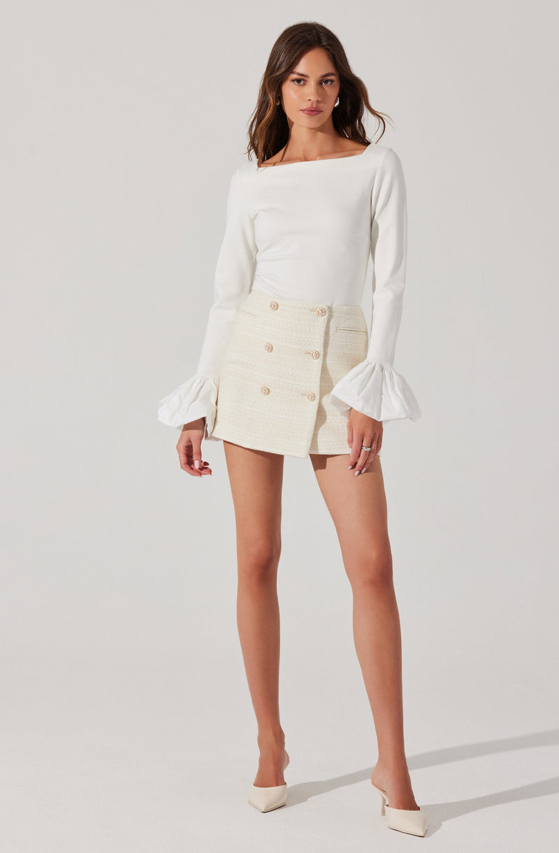 A woman is wearing a long-sleeved white top with a boat neckline and ruffled cuffs. She pairs it with a high-waisted, cream-colored, double-breasted mini skirt featuring gold buttons. She completes the look with white pointed high heels.