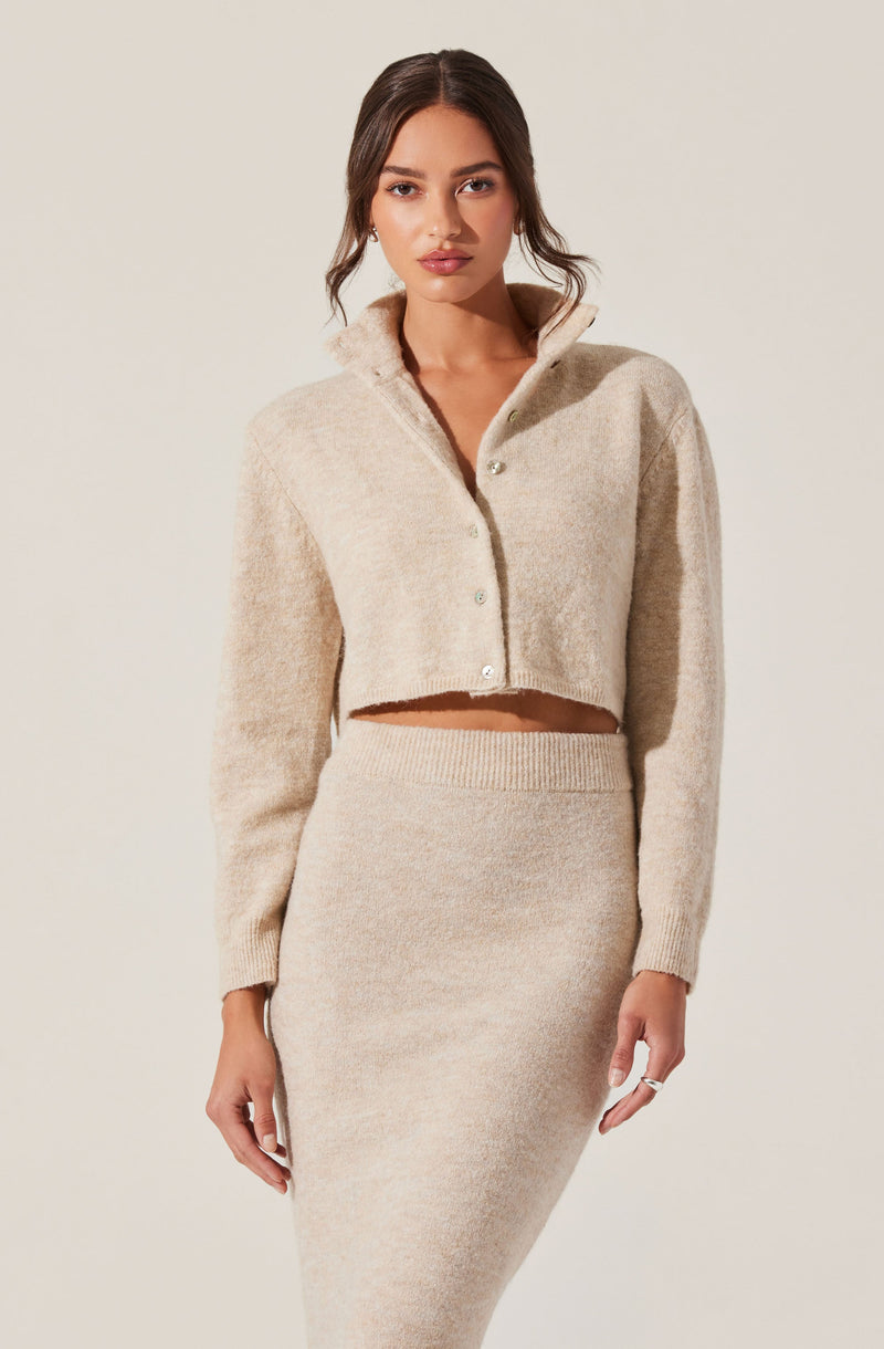 A woman is wearing a light beige, cropped, button-up cardigan with a high collar, paired with a matching high-waisted, fitted knit skirt.