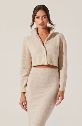 A woman is wearing a light beige, cropped, button-up cardigan with a high collar, paired with a matching high-waisted, fitted knit skirt.