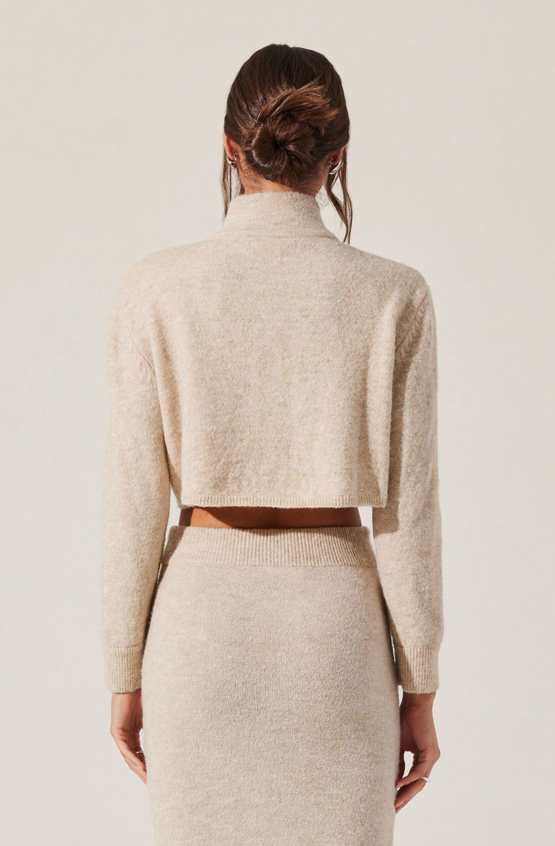 A woman is wearing a beige, cropped, long-sleeve sweater with a high neckline, paired with a matching high-waisted, fitted skirt. The sweater appears soft and slightly fuzzy, and the skirt is ribbed at the waistband. The woman's hair is styled in a loose bun with strands hanging down the sides.