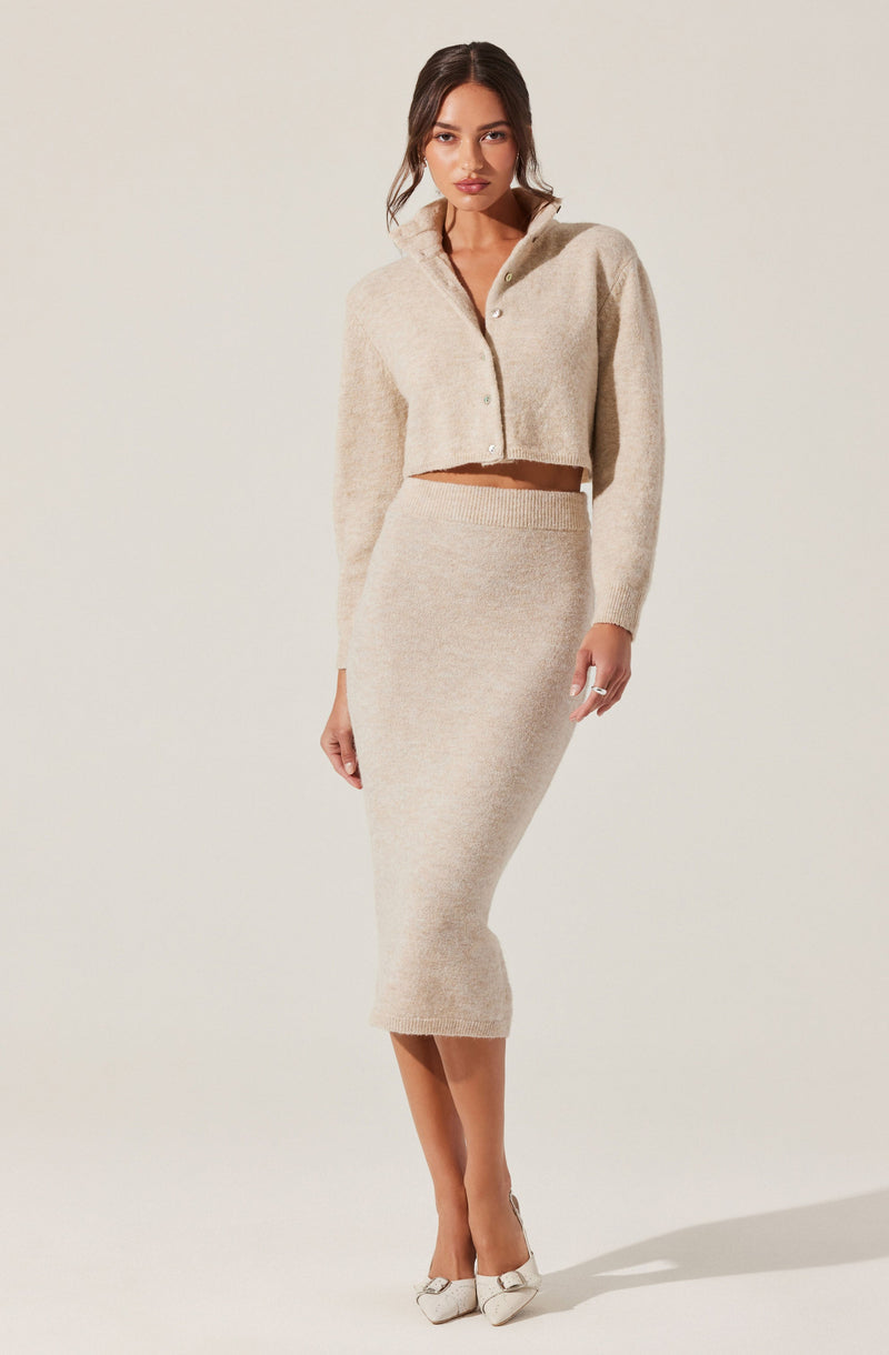 A woman is wearing a matching beige knit outfit consisting of a cropped, button-up sweater with a collar and a high-waisted, mid-length pencil skirt. She is also wearing white pointed-toe heels with decorative bows.
