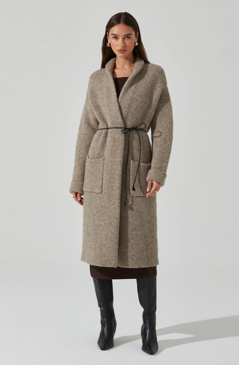 A woman is wearing a long, beige, textured coat made of a wool-like material, with a shawl collar and large front patch pockets. The coat is tied at the waist with a thin black drawstring, creating a cinched silhouette. Underneath, she has on a dark brown dress that reaches below the coat's hem. She completes her outfit with knee-high black leather boots that have a slight heel.