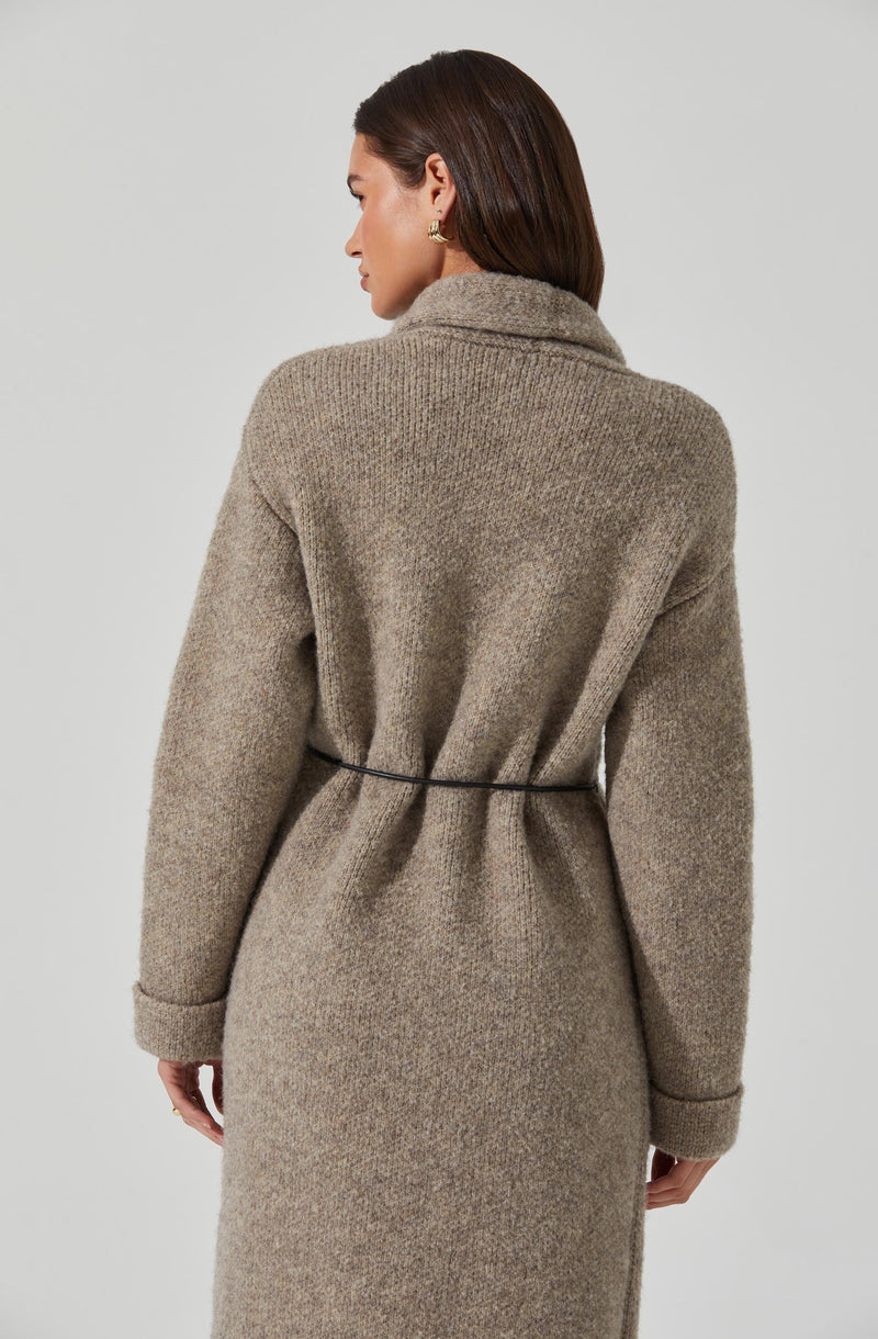 A woman is wearing a long, beige, textured knit coat with a high collar, and a thin black belt cinching the waist. The coat has a soft, slightly fuzzy appearance, with wide sleeves that are cuffed at the wrists.