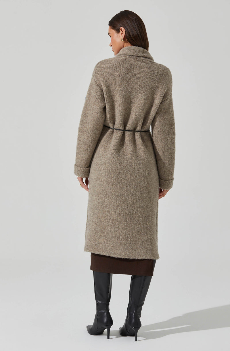 A woman is wearing a long, beige, woolen coat with a high collar and a belted waist. The coat extends below her knees, and she has on black high-heeled boots.