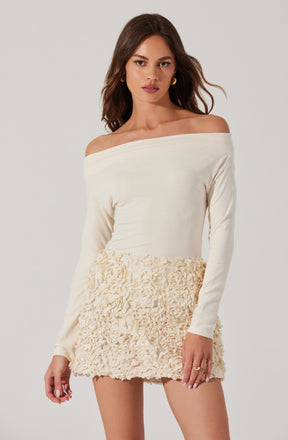 A woman is wearing an off-the-shoulder, long-sleeved, cream-colored top with a smooth, fitted fabric. She pairs it with a high-waisted, textured mini skirt that features a fluffy, ruffled floral pattern in a matching cream tone.