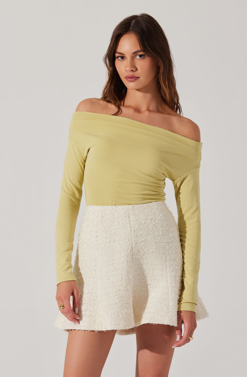 A woman is wearing a light yellow, off-the-shoulder long-sleeve top and a high-waisted cream-colored textured skirt with a frayed hem.