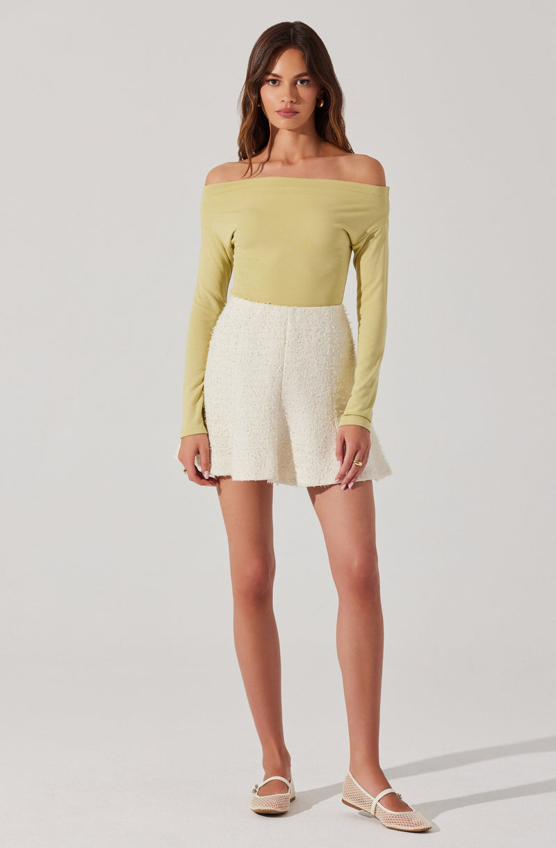 A woman is wearing a light yellow off-the-shoulder long-sleeve top paired with a cream-colored textured mini skirt. She is also wearing beige woven flat shoes with ankle straps.