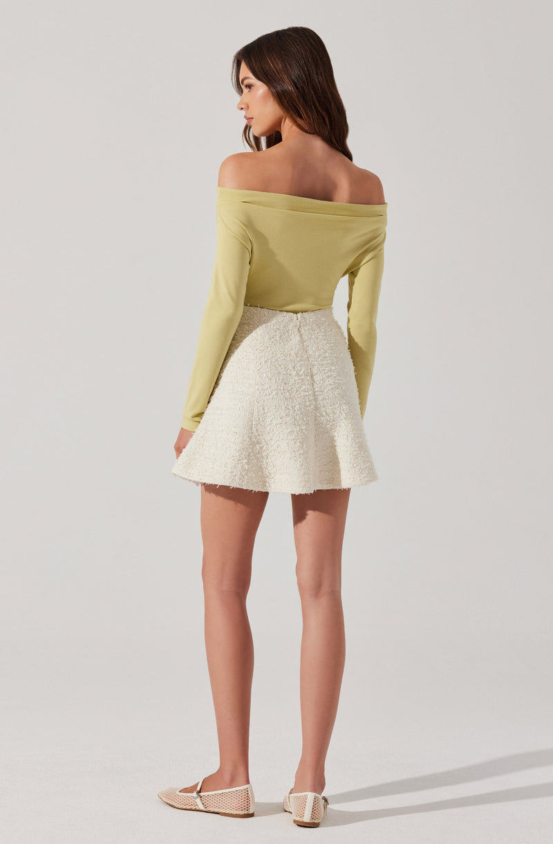 A woman is wearing an off-the-shoulder, long-sleeved, light yellow top paired with a high-waisted, white textured skirt that is flared and reaches mid-thigh. She is also wearing light-colored, woven flats.