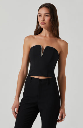 A young woman is wearing a strapless black crop top with a deep V neckline accented by a row of small, shiny embellishments along the edge. She pairs it with high-waisted black trousers.