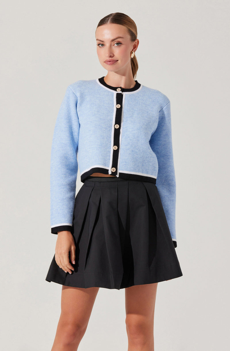 A woman is wearing a light blue cropped cardigan with black trim and large decorative buttons down the front. The cardigan has long sleeves, and the black trim is present along the collar, cuffs, and hem. She is also wearing a high-waisted black pleated skirt that reaches above her knees.