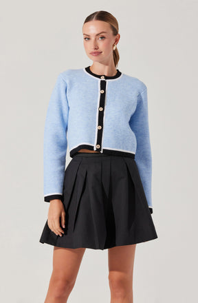 A woman is wearing a light blue cropped cardigan with black trim and large decorative buttons down the front. The cardigan has long sleeves, and the black trim is present along the collar, cuffs, and hem. She is also wearing a high-waisted black pleated skirt that reaches above her knees.