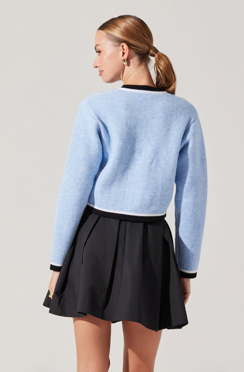 A woman is wearing a light blue, long-sleeved, cropped sweater with black and white trim at the cuffs and neckline, paired with a black pleated skirt that has a wide waistband and pleats.