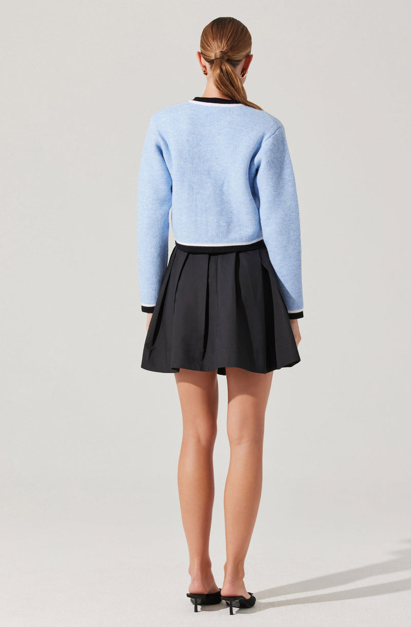 A person is wearing a light blue long-sleeve sweater with black and white trim at the collar, cuffs, and hem. They are also wearing a high-waisted black pleated skirt that falls above the knees, paired with black open-toe high heel shoes.