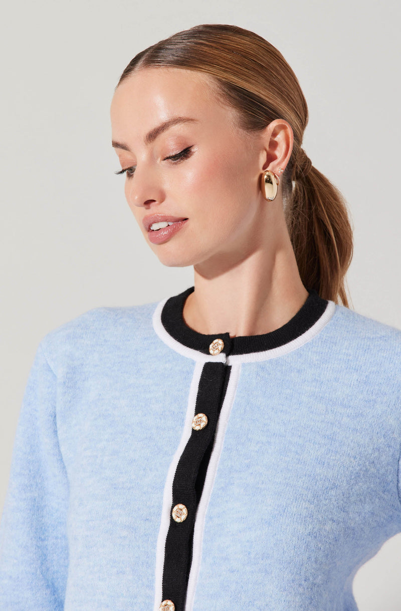 A woman with light skin and auburn hair styled in a low ponytail is wearing a light blue cardigan with black trim around the collar and down the front. The cardigan features white piping along the edges and decorative gold buttons. She is also wearing gold hoop earrings.