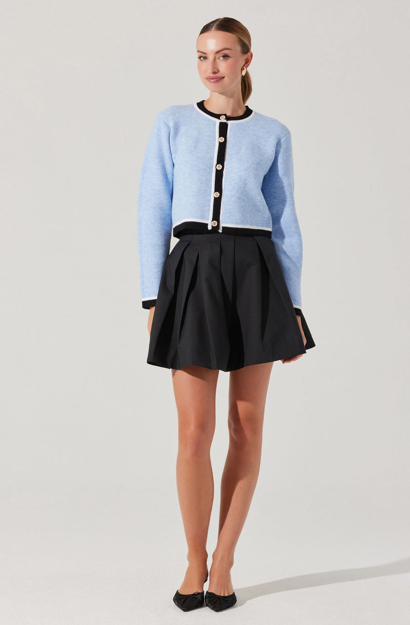 A young woman is wearing a light blue, cropped cardigan with black and white trim along the neckline, front opening, and cuffs. The cardigan has large, round buttons down the front. She pairs it with a high-waisted black pleated skirt that flares out slightly and ends above the knees. She is also wearing black pointed-toe flats.