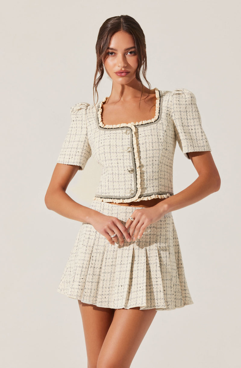 A woman is wearing a coordinated two-piece outfit made of a cream-colored, checkered textured fabric. The cropped top has short puffed sleeves and a square neckline, with decorative trim along the edges and down the front. The matching skirt is a high-waisted, pleated style that falls mid-thigh, also featuring the same checkered pattern and trim details.