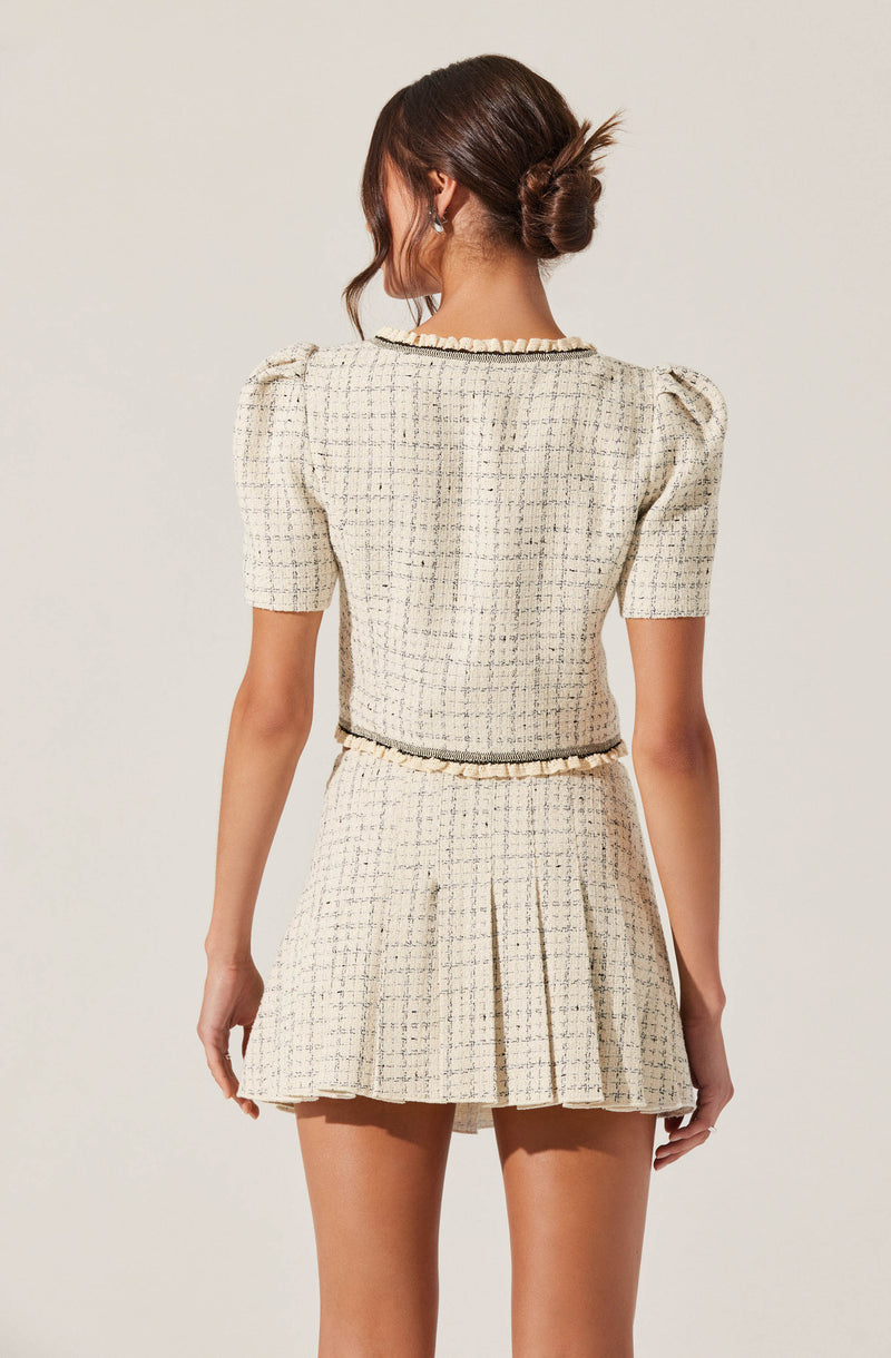 A woman is wearing a matching two-piece outfit made of a textured, cream-colored fabric with a subtle grid pattern. The top has short puffed sleeves and a round neckline with a ruffled trim. The skirt is high-waisted with a pleated design and a matching ruffled trim at the waist, creating a coordinated look. The outfit has a classic, elegant style with a touch of vintage inspiration.