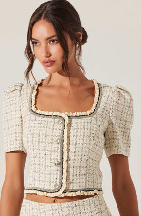 A woman is wearing a short-sleeved, cream-colored, textured tweed jacket with black trim along the neckline, front opening, and hem. The jacket features a rounded square neckline, puffed shoulders, and decorative pearl buttons.