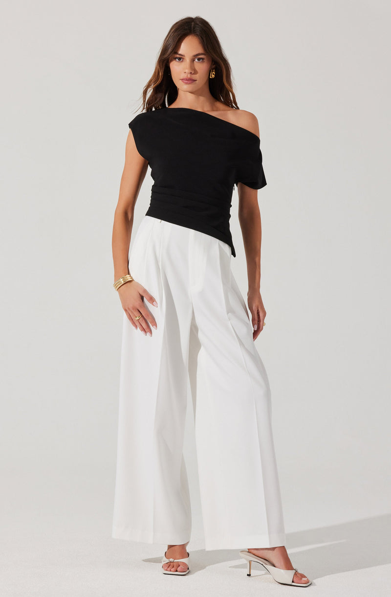 A woman is wearing an asymmetrical black top with a wide shoulder strap on one side and a sleeve on the other. She pairs it with high-waisted, wide-leg white trousers. She accessorizes with gold jewelry, including earrings, bracelets, and a ring. She also wears light-colored strappy heels.
