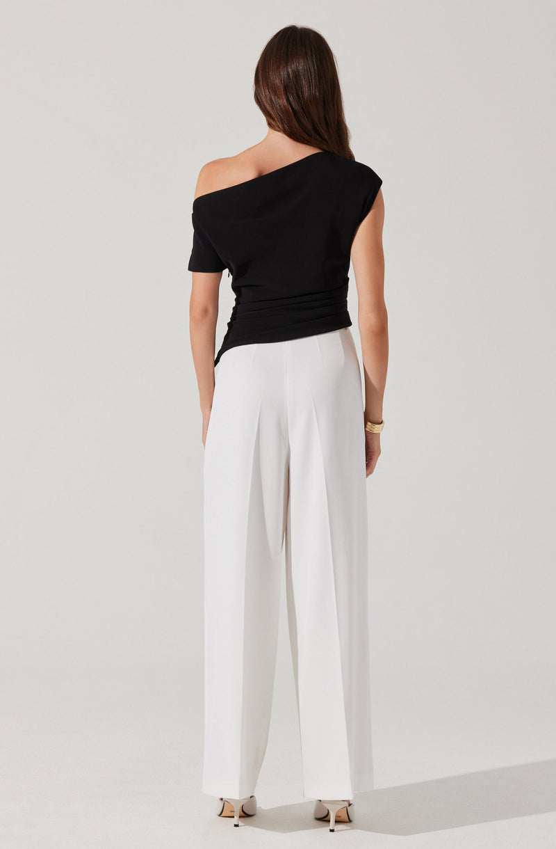 A woman is wearing a black, off-the-shoulder top with short sleeves and a relaxed fit. She pairs it with high-waisted, wide-leg white trousers that have pleats at the front. The outfit is complemented by a bracelet on her right wrist and beige high-heeled shoes.