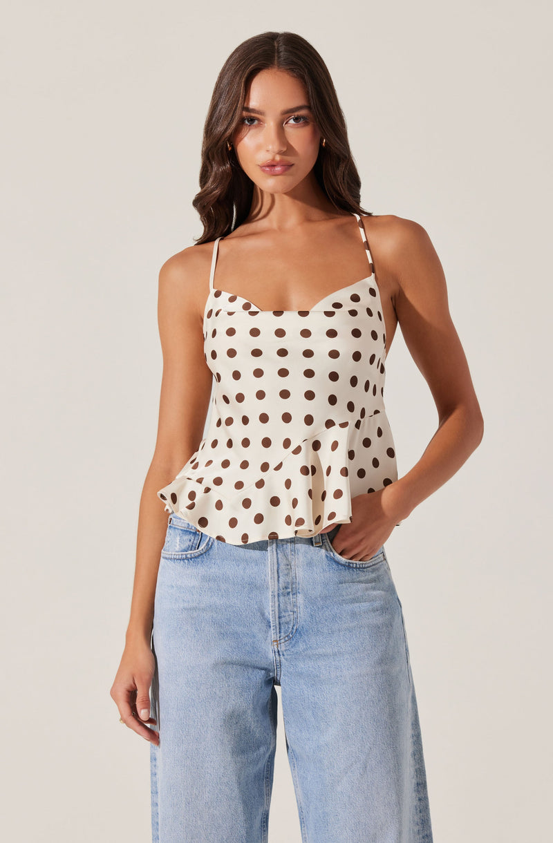 A young woman is wearing a sleeveless, cream-colored top with brown polka dots. The top has thin spaghetti straps, a sweetheart neckline, and a ruffled hem that flares slightly at the waist. She pairs it with light blue, high-waisted jeans.