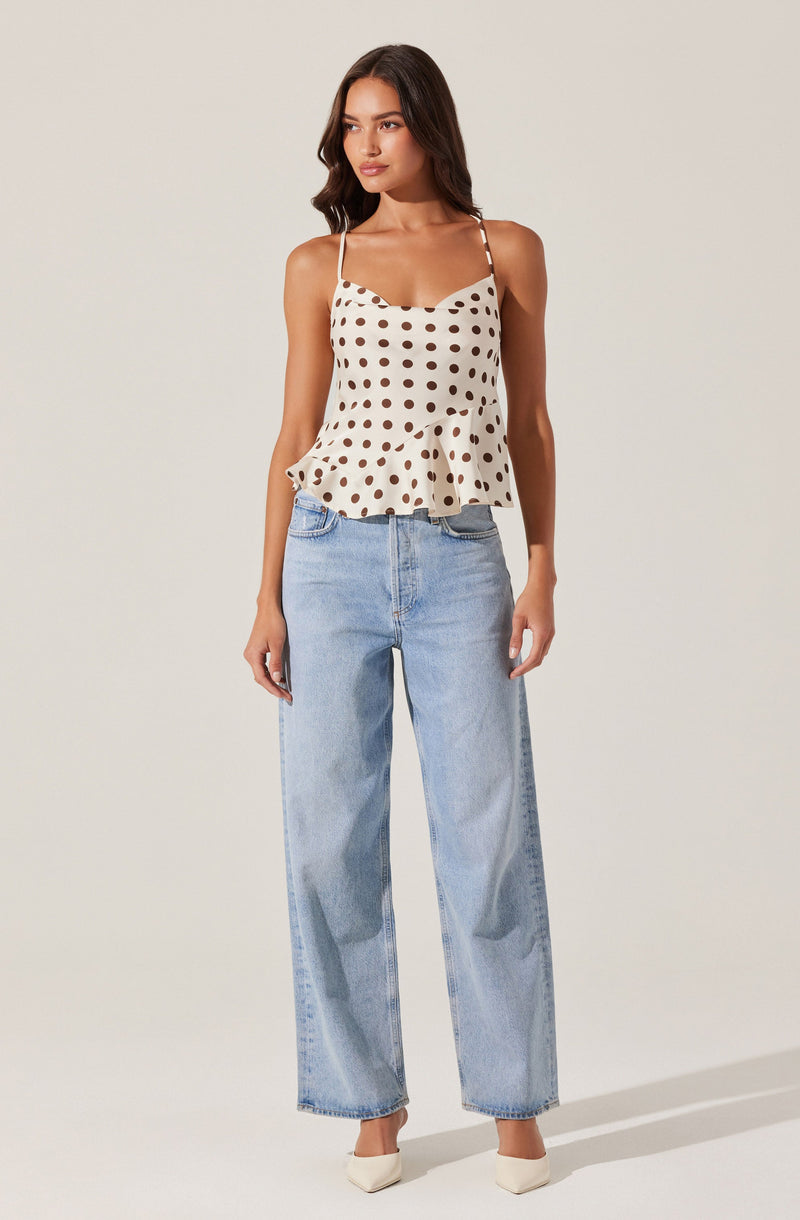 A woman is wearing a cream-colored sleeveless top with dark brown polka dots, featuring thin straps and a ruffled hem. She has paired it with light blue, high-waisted, wide-leg jeans. She is also wearing white pointed-toe shoes.