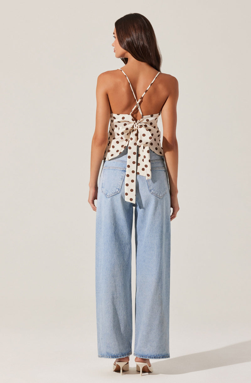 A woman is wearing a sleeveless, beige top with large brown polka dots and thin spaghetti straps that crisscross at the back and tie into a bow. She is also wearing high-waisted, light blue wide-leg jeans and beige heeled shoes.