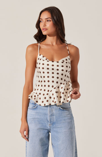 A woman is wearing a sleeveless, beige top with spaghetti straps and a dark brown polka dot pattern. The top features a ruffled hem and a slightly fitted bodice. She is also wearing high-waisted, light blue denim jeans.