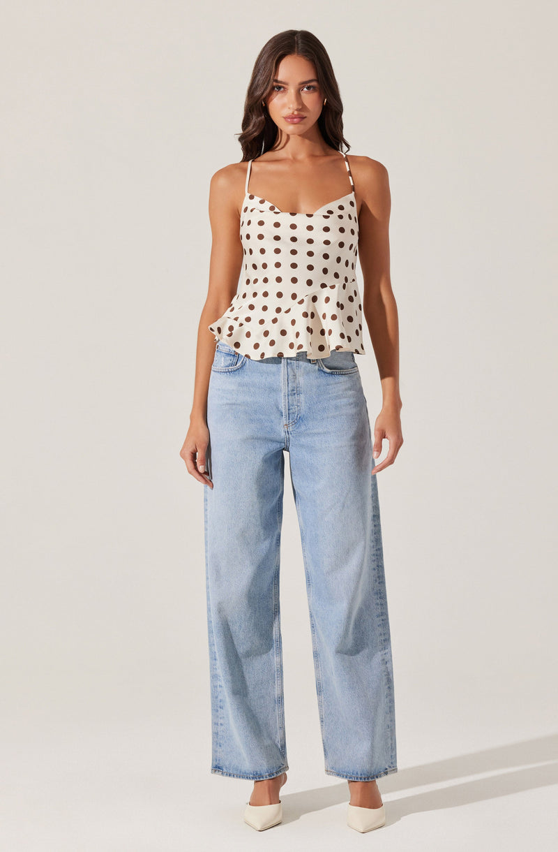 A woman is wearing a sleeveless, off-white top with large brown polka dots and thin spaghetti straps. The top has a ruffled hem that flares out slightly. She is also wearing high-waisted, light-wash wide-leg jeans and white pointed-toe shoes.